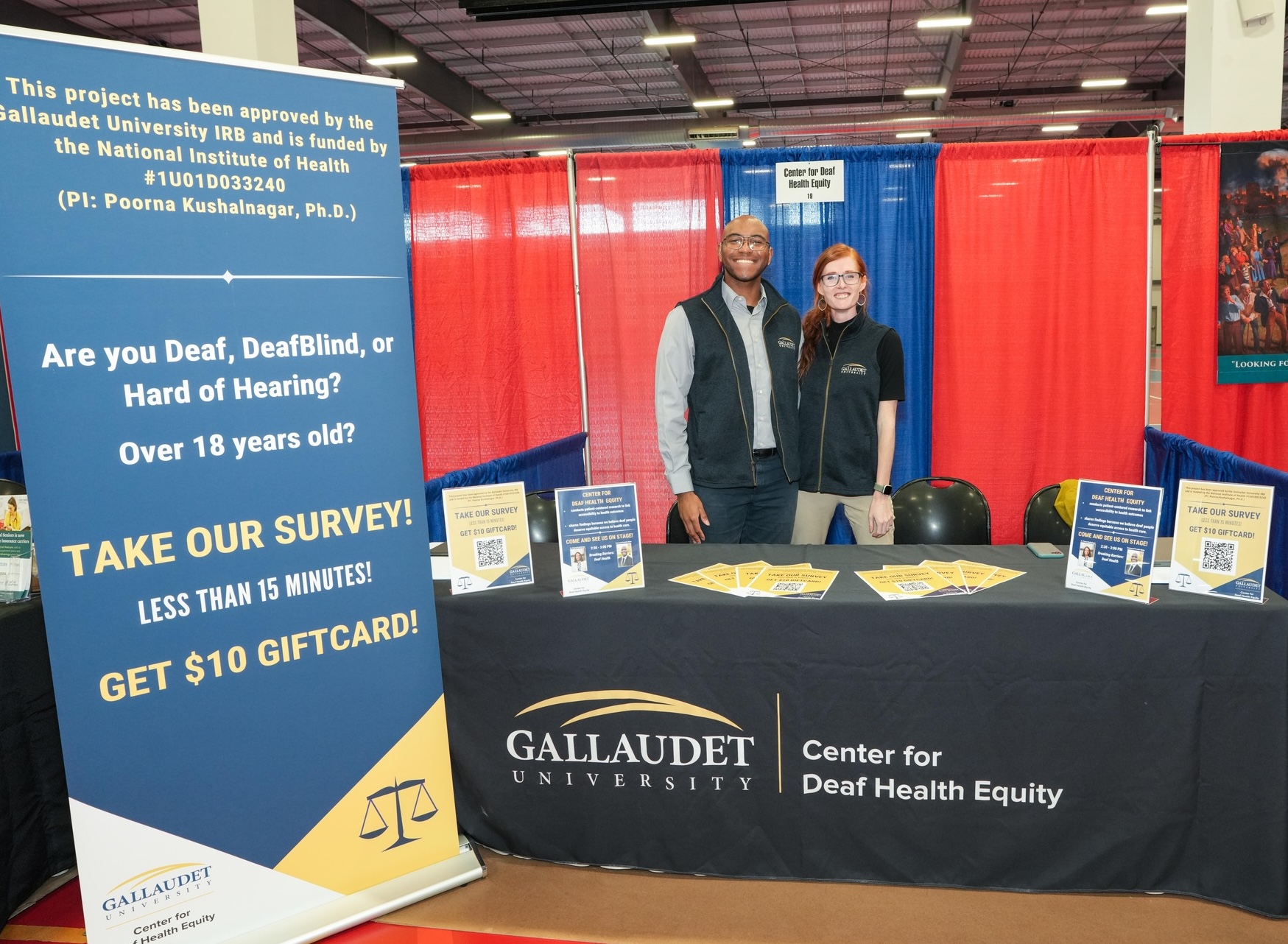 Emmanuel Perrodin-Njoku (a Black man in a gray suit and black vest) and Erika Bergeron (a white woman with red hair, wearing a black vest and short black shirt) smile while standing behind a booth table with a sign reading "Gallaudet University | Center for Deaf Health Equity."