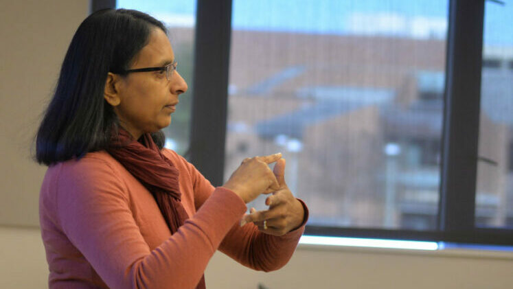 Dr. Poorna Kushalnagar, a South Asian woman with glasses, wearing a pink long-sleeved shirt and dark red scarf, stands presenting in front of a blurred classroom window in Gallaudet University's Hall Memorial Building.